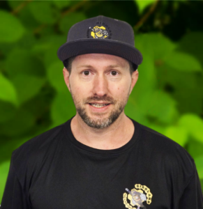 A man from the Richland team, sporting a beard, wears a black cap and a matching black t-shirt with a logo, standing confidently in front of a green, leafy background.