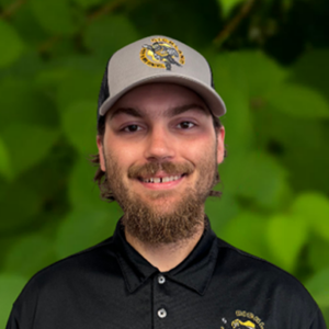 A man with a beard and baseball cap smiles at the camera. He is wearing a black collared shirt with a Richland team emblem on the left side. The background is a blurred green foliage.