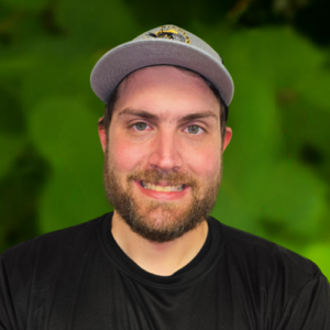 A man from the Richland team smiles at the camera, sporting a beard and a cap, dressed in a black shirt against a green blurred background.