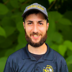 A man with a beard wears a gray cap and a dark blue polo shirt adorned with the Richland Team logo, smiling warmly against a leafy green backdrop.