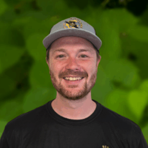 A man wearing a gray cap and black t-shirt smiles at the camera, representing the Richland team, with a blurred green leafy background behind him.