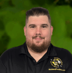 A man with short dark hair and a beard, part of the Richland team, wears a black polo shirt with a yellow bee logo and "RichlandParishSo.com" on the chest, posed in front of a green background.