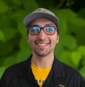 A member of the Richland team wearing glasses, a gray cap, and a black jacket over a yellow shirt smiles in front of a leafy green background.
