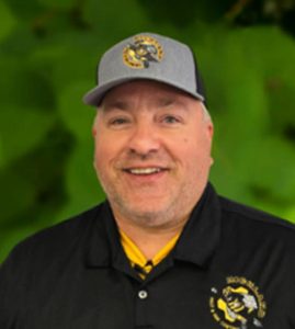 A man wearing a gray cap and black polo shirt with a bee logo, representing the Richland team, stands in front of a blurred green background.