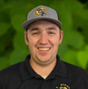 A man wearing a gray cap and black collared shirt stands smiling in front of a green leafy background, representing the Richland team.