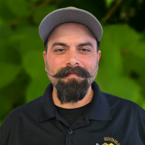 Man with a styled mustache and beard, wearing a gray cap and black collared shirt with a yellow and white logo, representing the Richland team, standing in front of a green background.