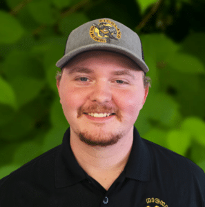 A man wearing a gray hat and black polo shirt with a circular Richland Team logo, smiling in front of a leafy green background.