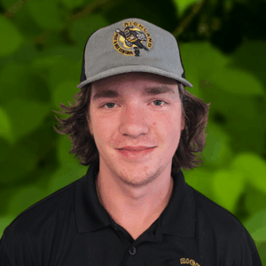 Young man wearing a gray cap and black polo shirt with a yellow and black Richland Team logo, standing in front of a green leafy background.
